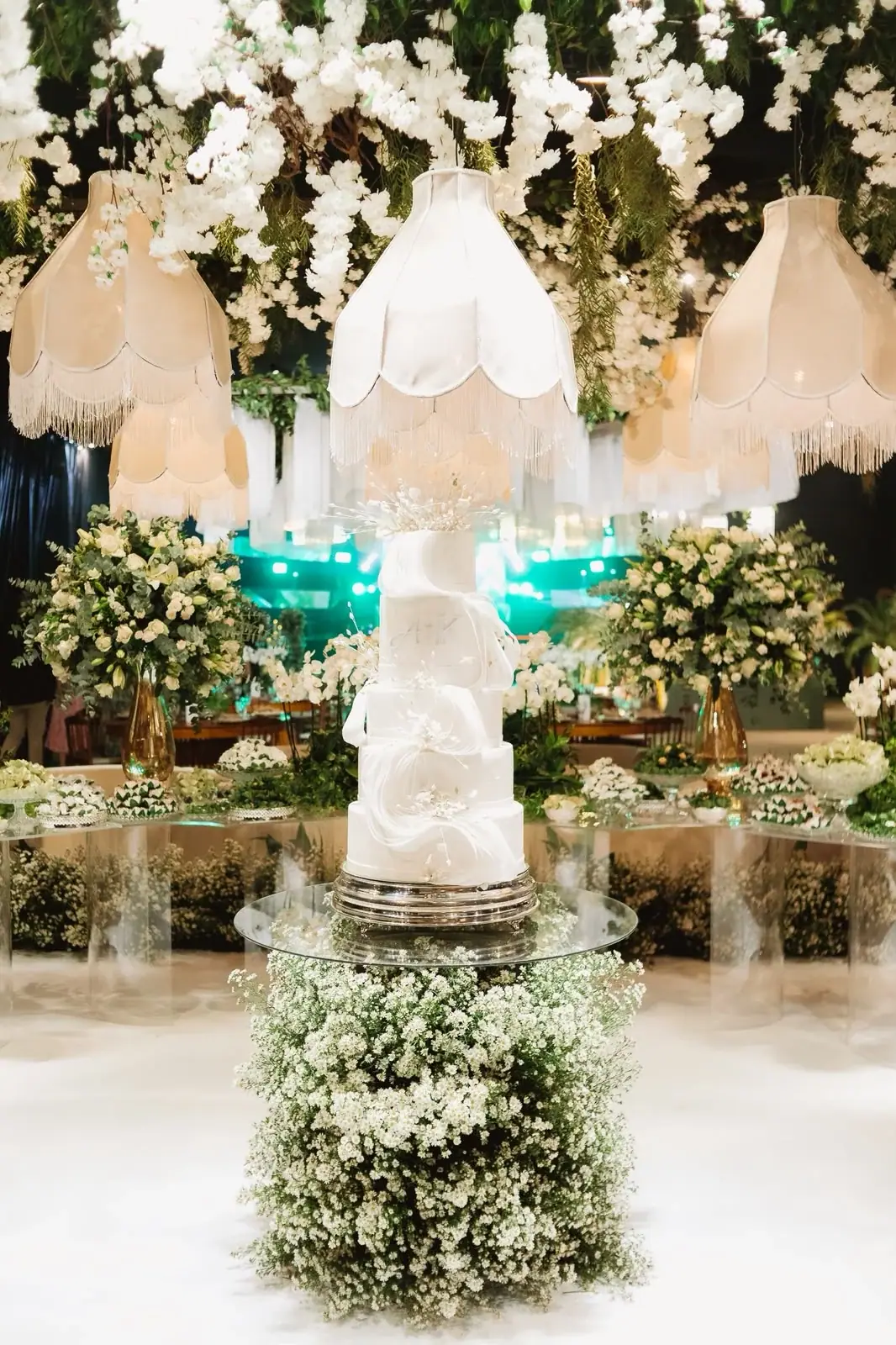 Bolo branco sobre pedestal de flores, com luminárias de tecido e arranjos ao redor.